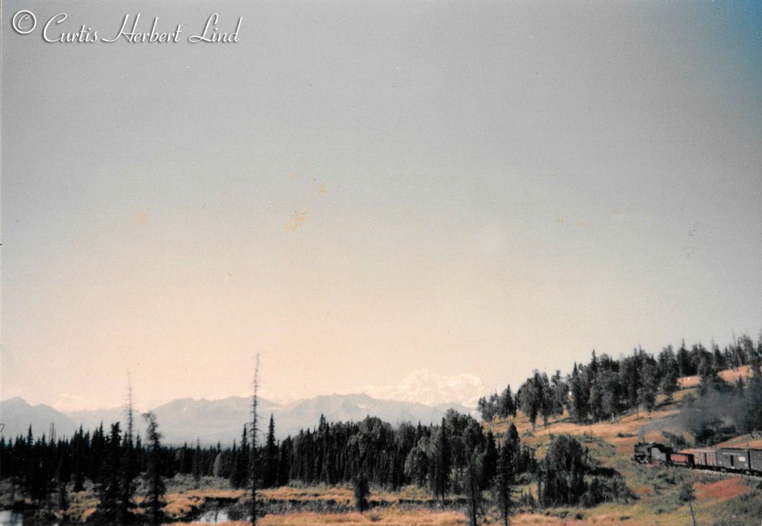 Look closely or you will miss the subject of interest in this expanse of late fall scenery. That is a 400 class Consolidation pulling a pretty good string of cars, assuming the photographer is on the same train. Looks like a Hart Convertible Gon, an outside braced box car and at least two Pullman troop sleepers still in OD paint. Between Chulitna and Hurricane.  Mt. McKinley is faintly showing in the background.